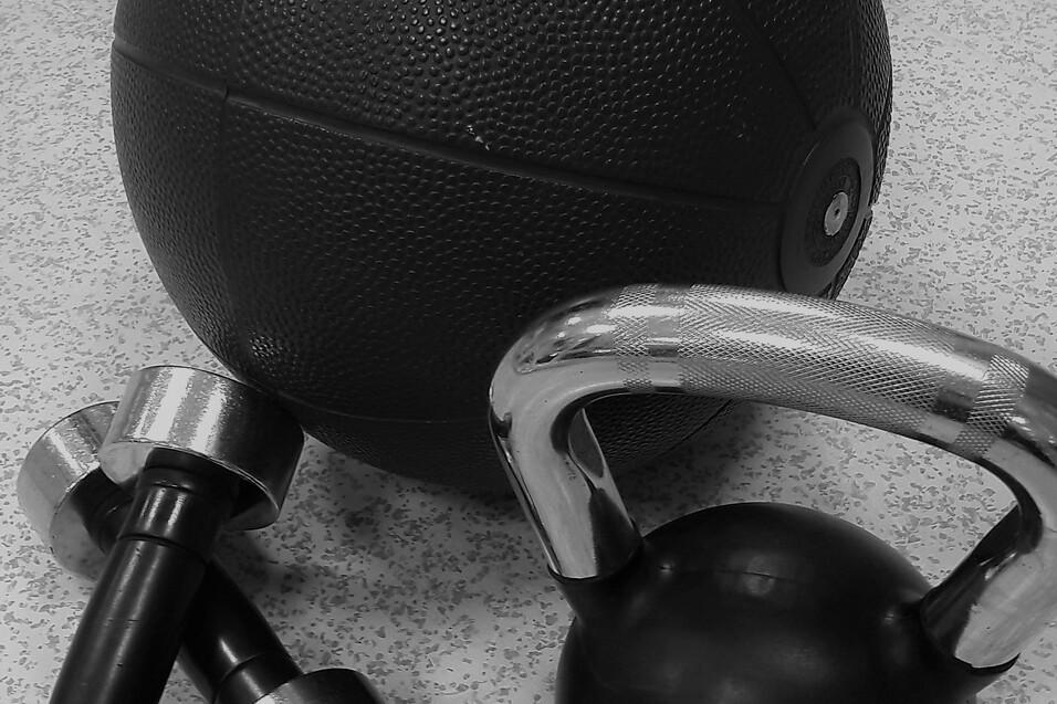 Nahaufnahme eines Gymnastikballs, einer Kettlebell und einer Hantel auf einem grauen Boden.