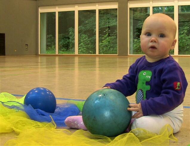 Ein kleines Kind sitzt auf dem Boden einer Sporthalle und spielt mit bunten Bällen und Tüchern.