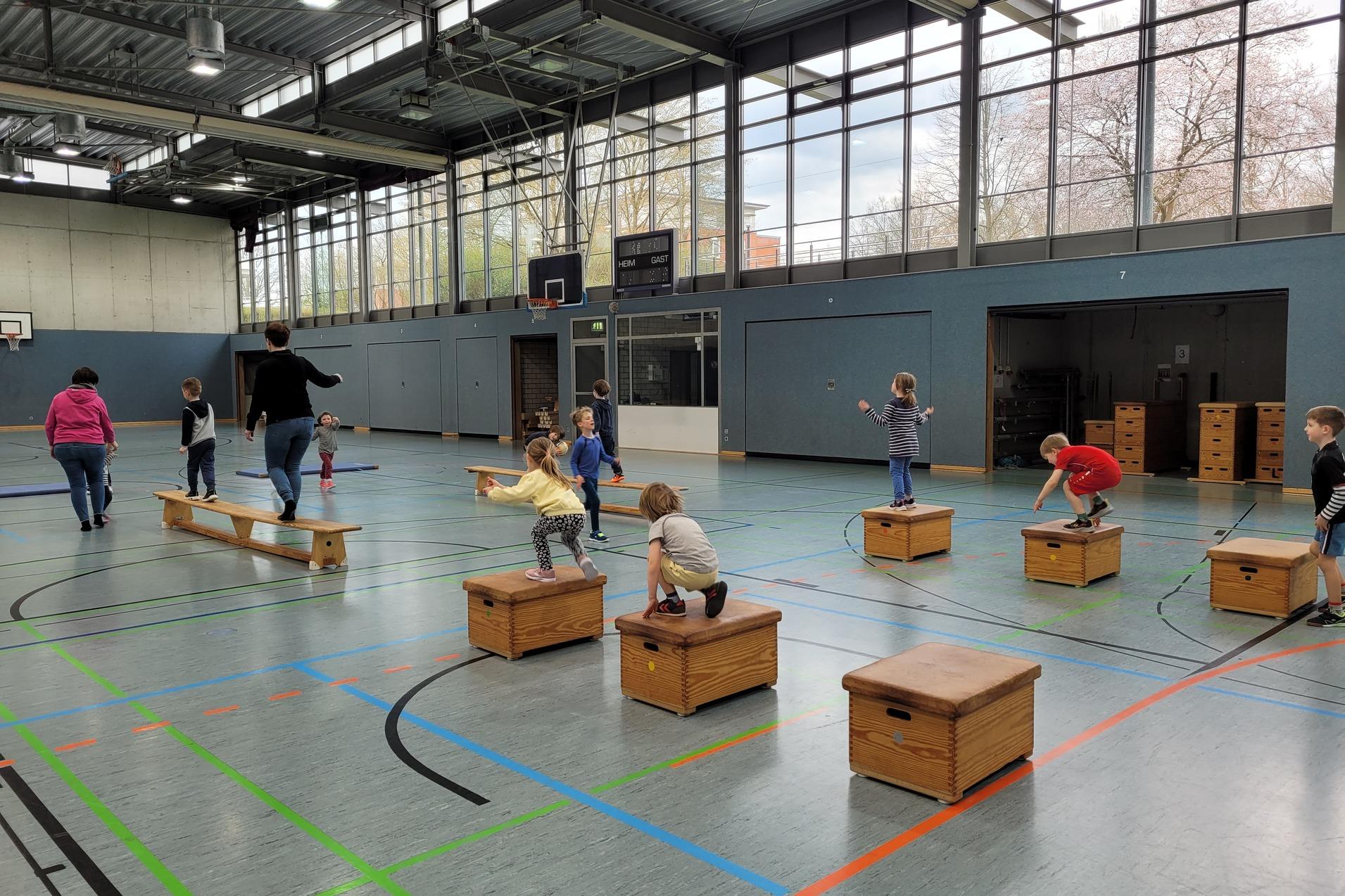 Kinder und Erwachsene &uuml;ben Balancieren und Springen auf Holzk&auml;sten in einer Sporthalle mit gro&szlig;en Fenstern.
