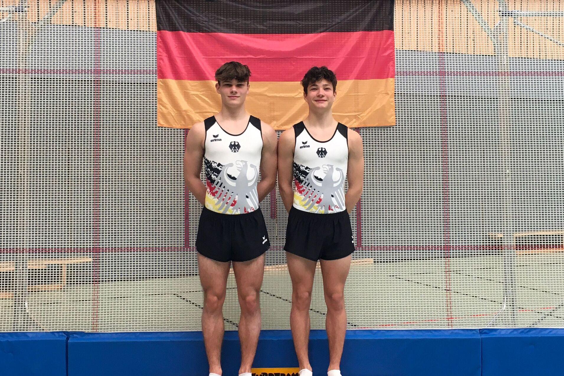 Zwei Turner in Trikots mit deutschem Adler posieren auf einer Trainingsmatte vor einer Deutschlandflagge.