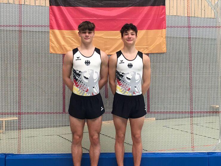 Zwei Turner in Trikots mit deutschem Adler posieren auf einer Trainingsmatte vor einer Deutschlandflagge.