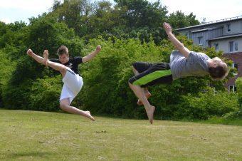 Zwei Jugendliche führen im Sprung Karate-Elemente auf einer Wiese vor, umgeben von Bäumen und Gebäuden.