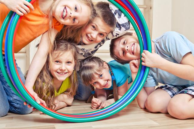 Gruppe von fünf lachenden Kindern, die durch bunte Reifen schauen und auf einem Holzboden sitzen.
