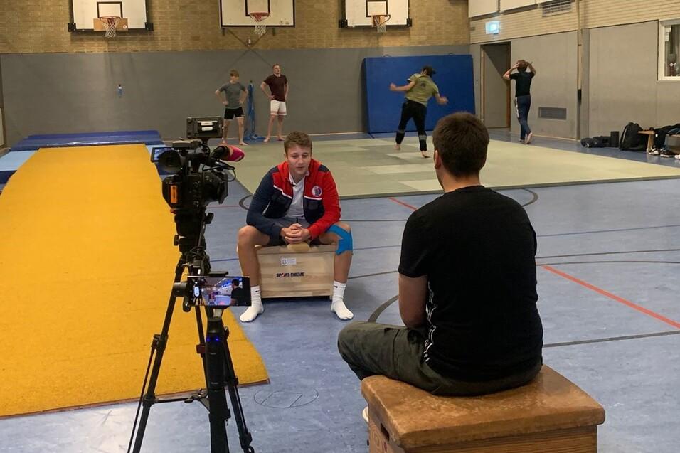 Training in a sports hall with a camera focused on a seated athlete and others practicing in the background.