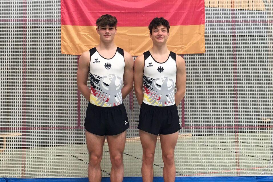 Zwei Turner in Trikots mit deutschem Adler posieren auf einer Trainingsmatte vor einer Deutschlandflagge.