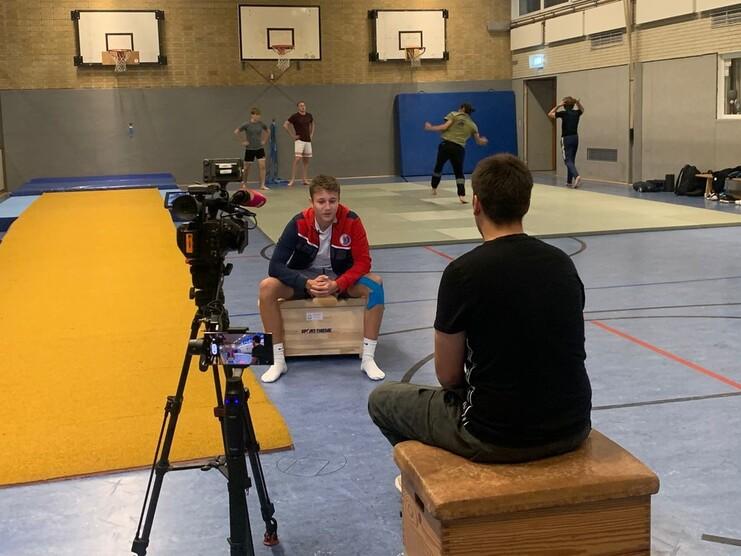 Training in a sports hall with a camera focused on a seated athlete and others practicing in the background.