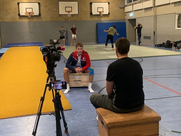 Training in a sports hall with a camera focused on a seated athlete and others practicing in the background.