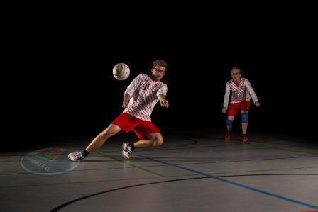 Zwei Männer spielen Volleyball in einem dunklen Raum, einer springt zum Ball, der andere beobachtet.