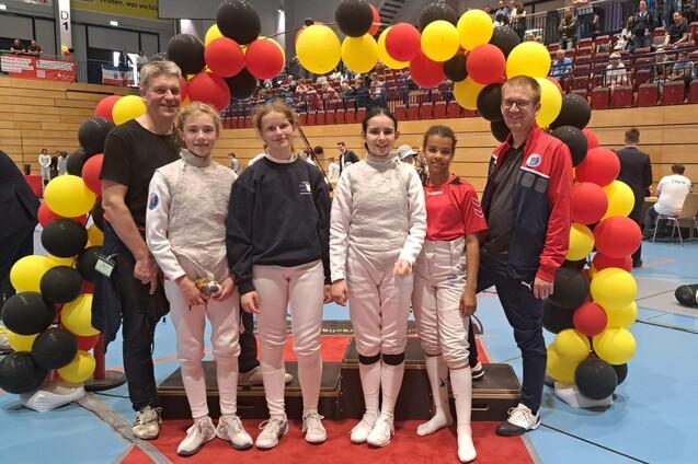 F&uuml;nf Personen, darunter drei in Fechtanz&uuml;gen, posieren vor einem bunten Luftballon-Bogen in einer Sporthalle.