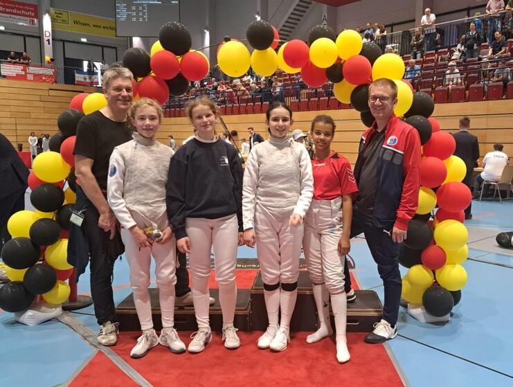 Fünf Personen, darunter drei in Fechtanzügen, posieren vor einem bunten Luftballon-Bogen in einer Sporthalle.