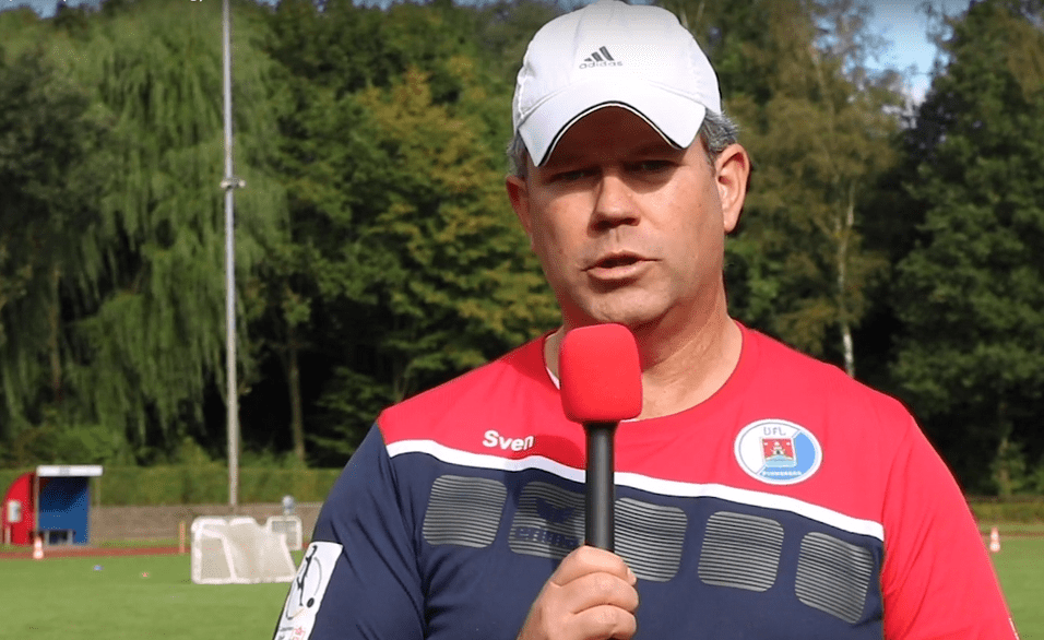 Mann mit Mikrofon spricht auf einem Fu&szlig;ballfeld, umgeben von B&auml;umen und Toren im Hintergrund.