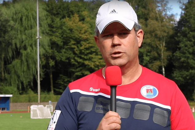 Mann mit Mikrofon spricht auf einem Fu&szlig;ballfeld, umgeben von B&auml;umen und Toren im Hintergrund.