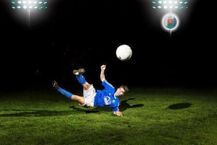Fußballspieler in blauer Uniform macht einen Fallrückzieher auf einem Rasenplatz unter Flutlicht.