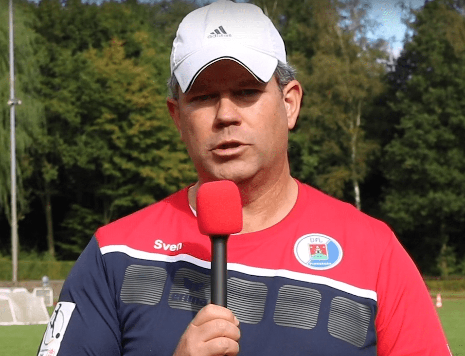 Mann mit Mikrofon spricht auf einem Fußballfeld, umgeben von Bäumen und Toren im Hintergrund.