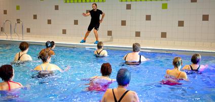 Wassergymnastik-Kurs mit einer Gruppe von Frauen im Pool, angeleitet von einem Trainer auf dem Beckenrand.