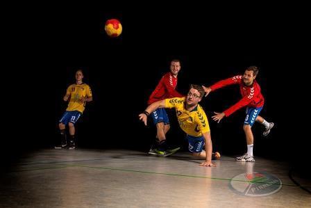 Vier Handballspieler in Aktion: Ein Spieler in Gelb springt nach einem Ball, während zwei in Rot und einer in Blau zusehen.