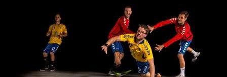 Vier Handballspieler in Aktion: Ein Spieler in Gelb springt nach einem Ball, während zwei in Rot und einer in Blau zusehen.
