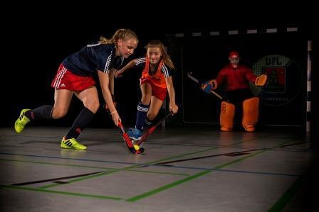 Zwei Hockeyspielerinnen kämpfen um den Ball in einer Halle, während ein Torwart im Hintergrund bereit ist.