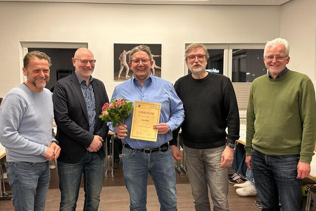 Gruppenfoto von f&uuml;nf M&auml;nnern, einer h&auml;lt eine Urkunde und einen Blumenstrau&szlig;, in einem Raum mit Tischen.