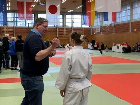 Ein Trainer spricht mit einem jungen Judoka in einem Sportzentrum, umgeben von Flaggen und anderen Wettkämpfern.