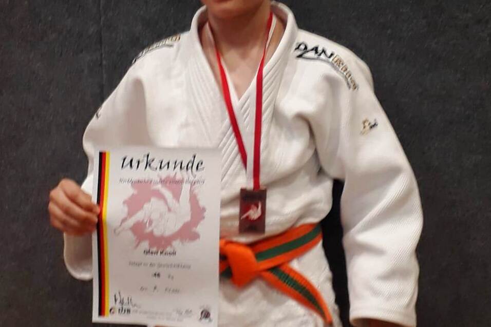 Ein junger Judoka in wei&szlig;em Gi h&auml;lt stolz eine Urkunde und eine Medaille vor einem grauen Hintergrund.