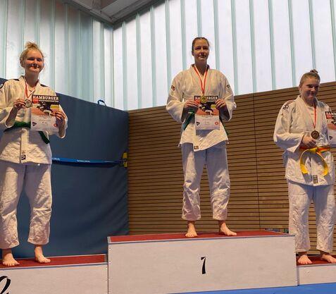 Drei Judokas stehen auf dem Siegertreppchen, halten Urkunden und tragen Medaillen in einer Sporthalle.