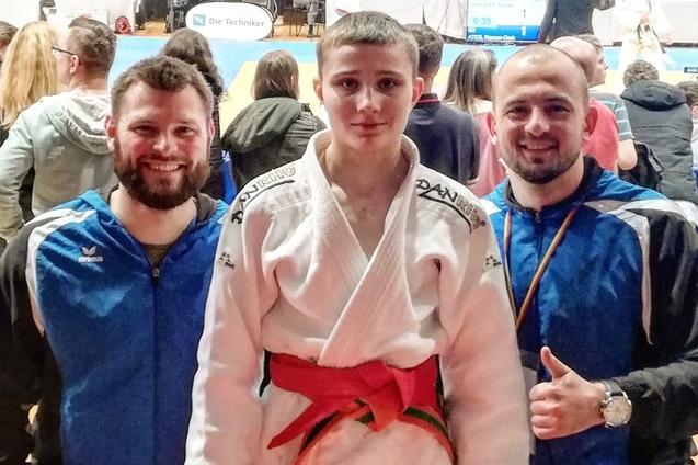 Drei Personen in Sportbekleidung stehen vor einer Wettkampfst&auml;tte mit Judo-Teppich und Zuschauern im Hintergrund.