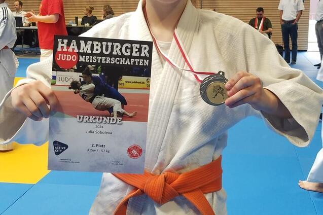 L&auml;chelndes M&auml;dchen in Judogi mit orangefarbenem G&uuml;rtel, h&auml;lt eine Urkunde und Medaille der Hamburger Judo Meisterschaften.