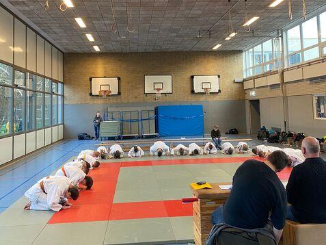 Judo-Training mit Teilnehmern in weißen Anzügen, die sich auf der Matte verbeugen, im Hintergrund Zuschauer und eine Sporthalle.