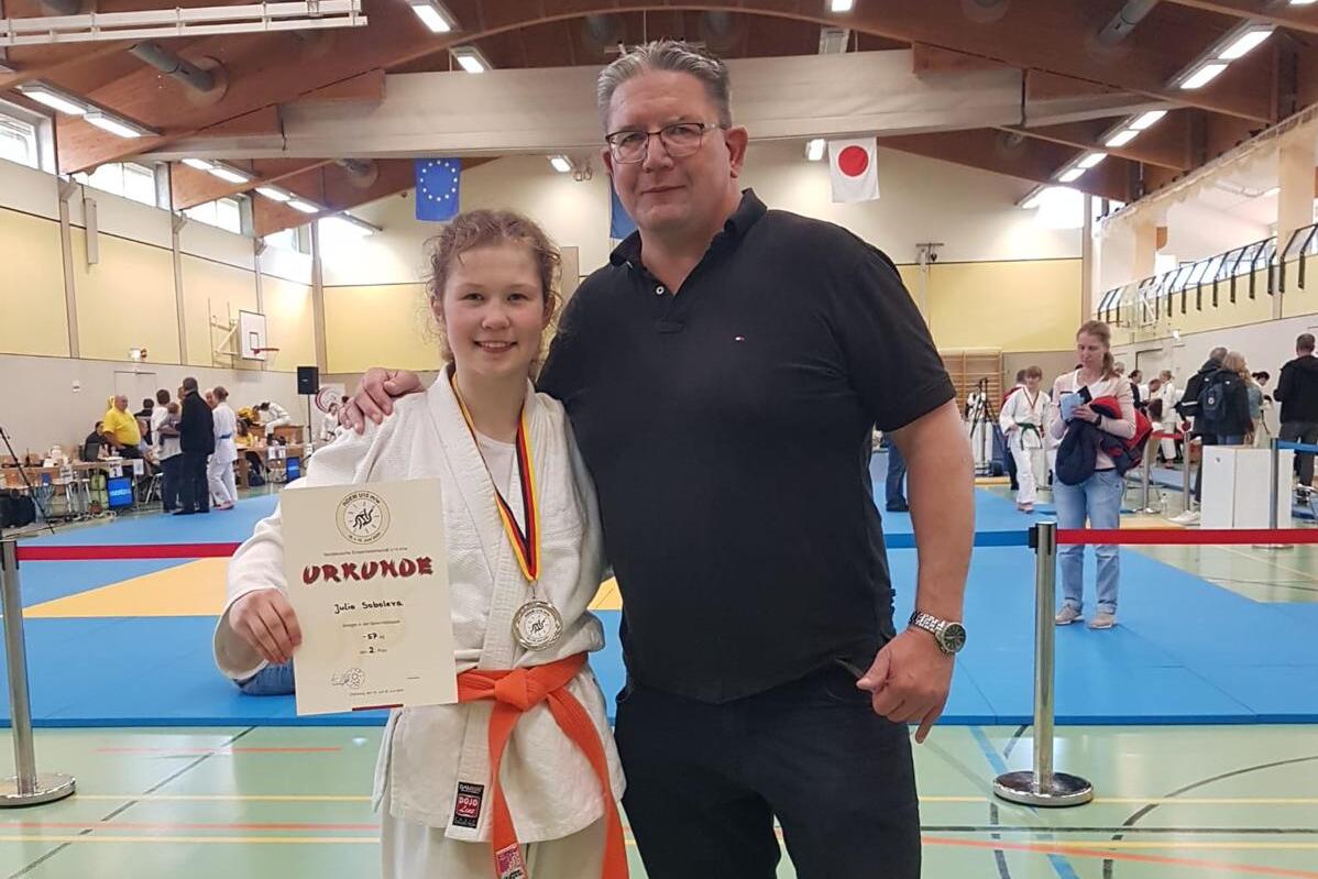 Junge in Karateanzug mit Medaille und Urkunde steht neben einem Mann in Sportbekleidung in einer Sporthalle.