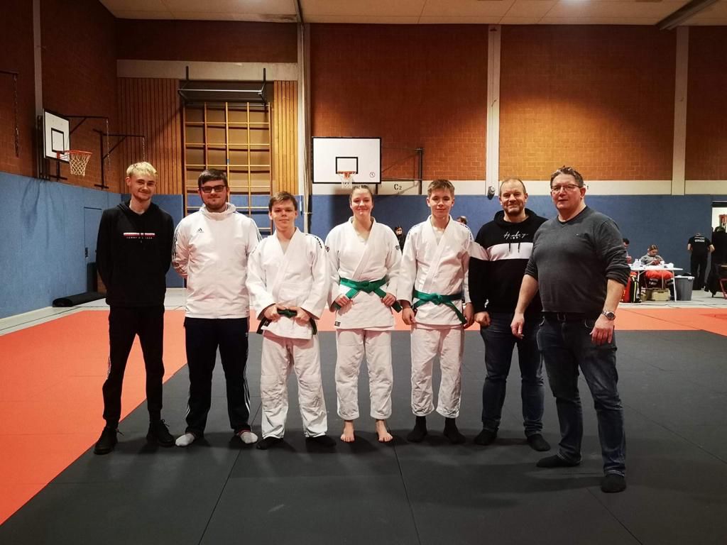 Gruppe von sechs Personen auf einer Judomatte; drei tragen Judoanzüge, zwei haben grüne Gürtel.