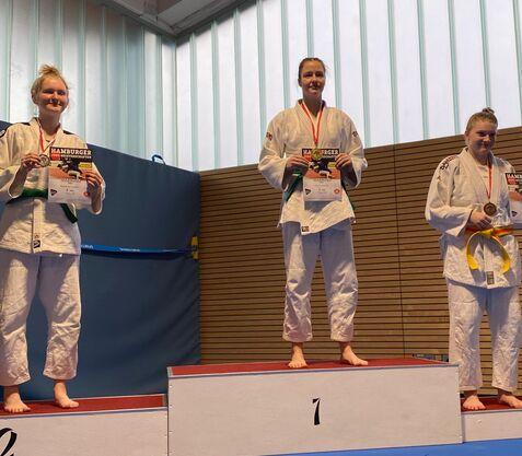 Drei Judokas stehen auf dem Siegertreppchen, halten Urkunden und tragen Medaillen in einer Sporthalle.
