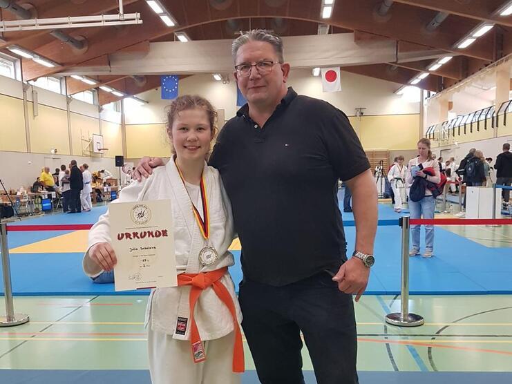 Junge in Karateanzug mit Medaille und Urkunde steht neben einem Mann in Sportbekleidung in einer Sporthalle.