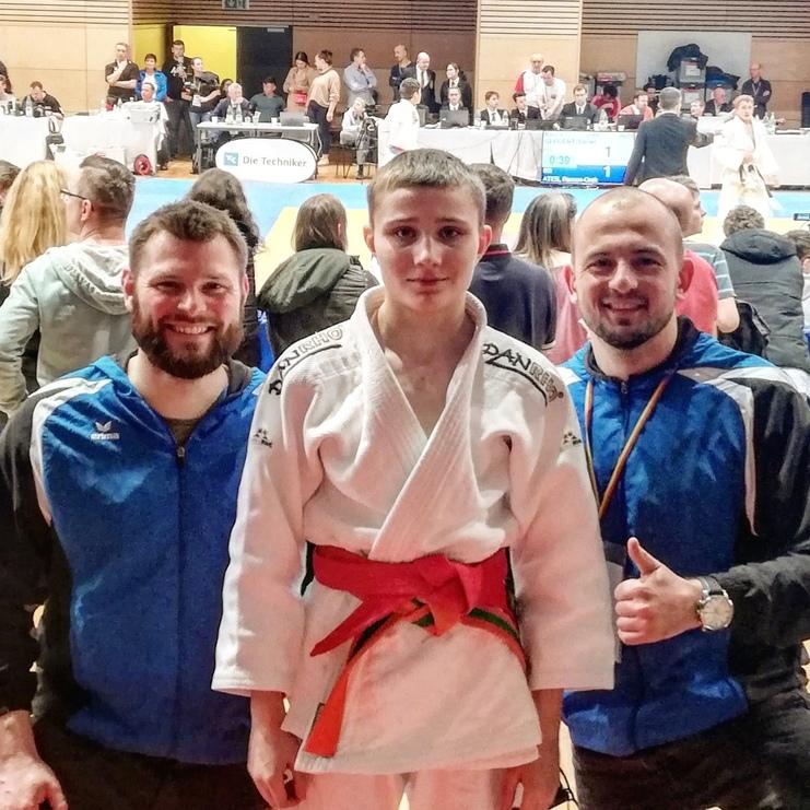 Drei Personen in Sportbekleidung stehen vor einer Wettkampfstätte mit Judo-Teppich und Zuschauern im Hintergrund.