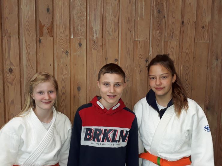 Drei Jugendliche stehen vor einer Holzwand; zwei tragen Judogis, der dritte ein rotes Sweatshirt.