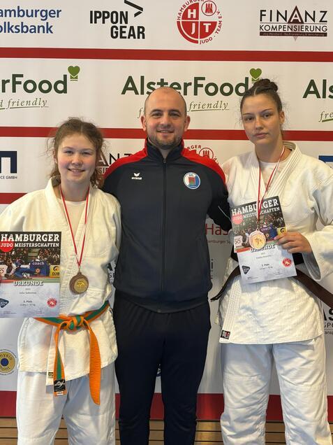 Zwei judosportliche Medaillengewinnerinnen mit ihren Urkunden stehen neben einem Trainer vor einem Werbebanner.