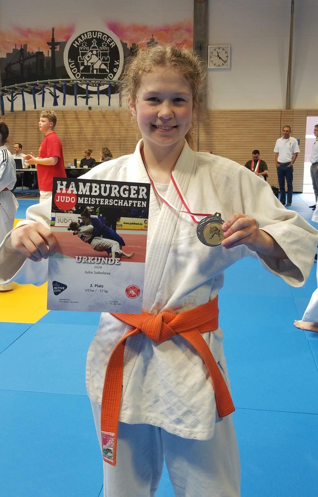 Lächelndes Mädchen in Judogi mit orangefarbenem Gürtel, hält eine Urkunde und Medaille der Hamburger Judo Meisterschaften.