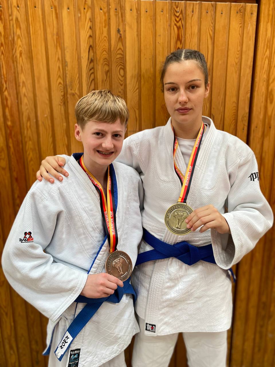 Zwei Judokas in Judokleidung stehen nebeneinander und zeigen stolz ihre Medaillen vor einer Holzwand.