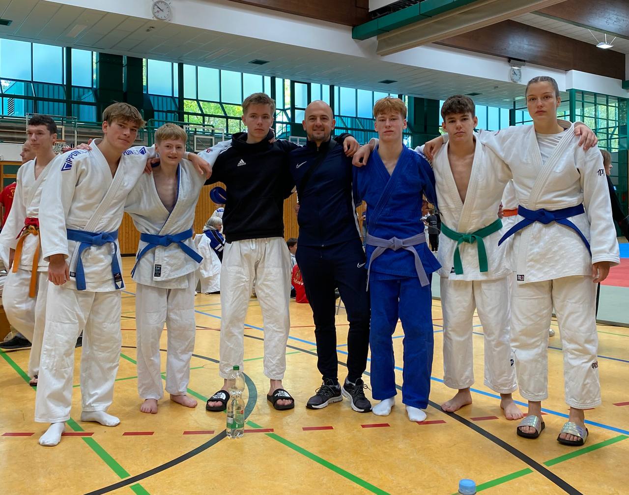 Gruppe von Judo-Athleten in ihren Anzügen mit Trainer in sportlicher Kleidung in einer Turnhalle.