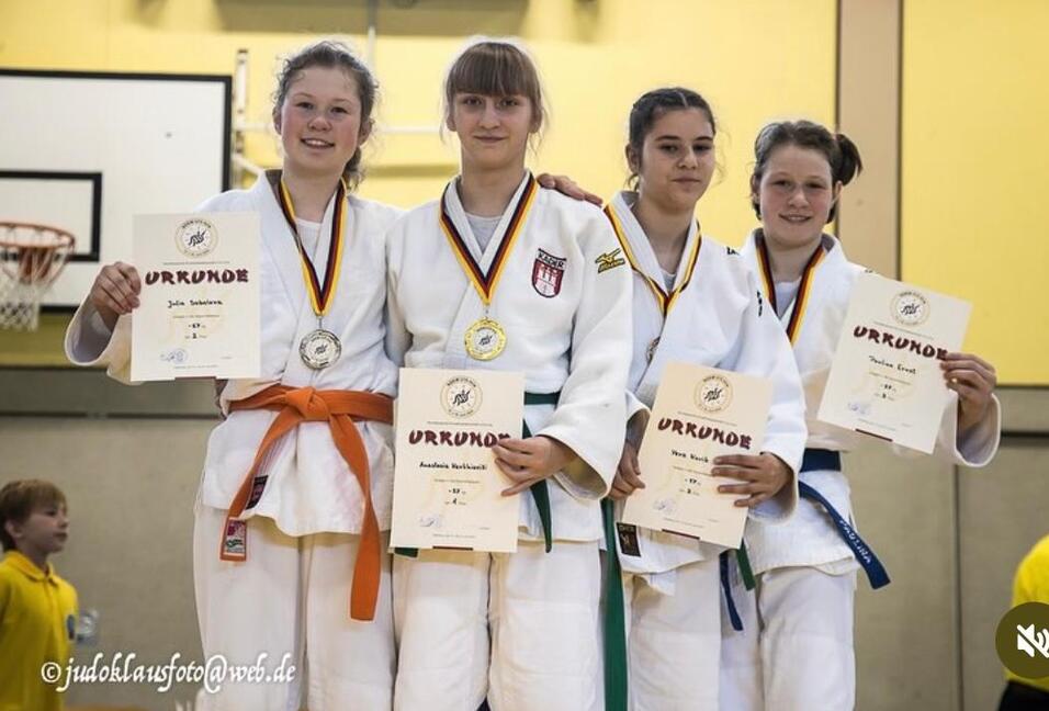 Vier Judoka in weißen Anzügen halten Urkunden und Medaillen, stehen vor einer Sporthalle mit Basketballkorb im Hintergrund.