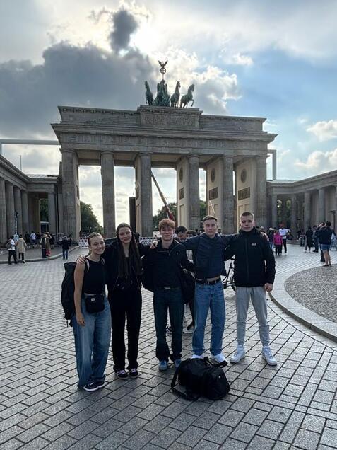 Gruppe von fünf Personen posiert vor dem Brandenburger Tor, umgeben von Besuchern auf einem gepflasterten Platz.