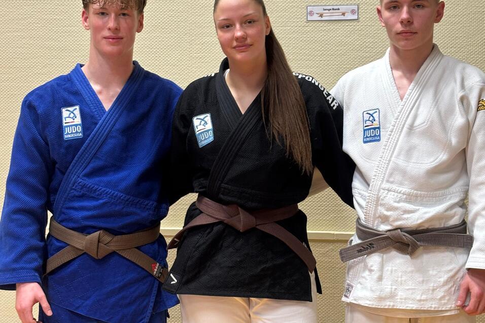 Drei Judokas stehen nebeneinander: ein Junge in blauem, ein M&auml;dchen in schwarzem und ein Junge in wei&szlig;em Judoanzug.