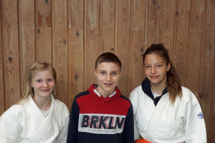 Drei Jugendliche stehen vor einer Holzwand; zwei tragen Judogis, der dritte ein rotes Sweatshirt.