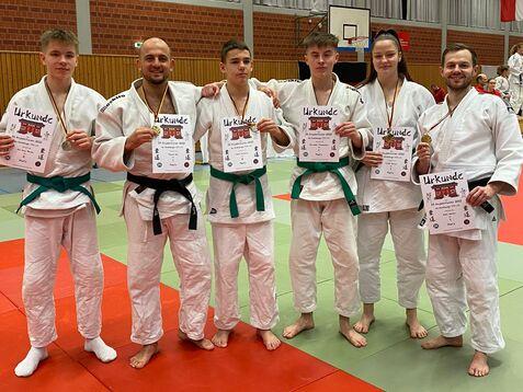 Sechsergruppe in Judokleidung h&auml;lt Urkunden; Hintergrund mit Landesflaggen in einer Sporthalle.