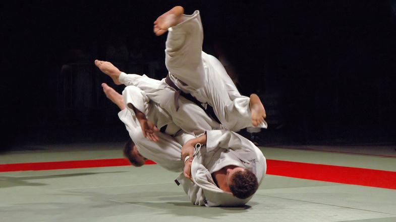 Zwei Judoka in weißen Anzügen führen einen Wurf auf einer Tatami mit rotem Rand aus.