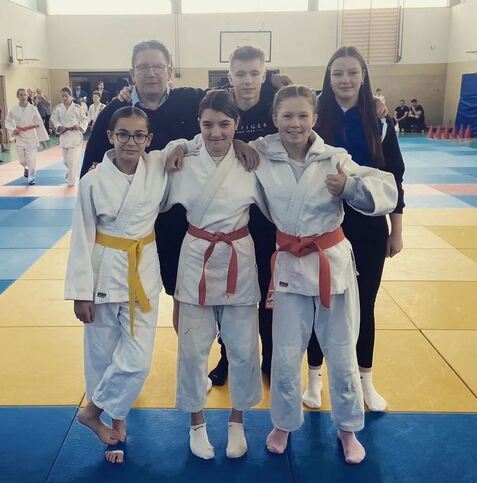 Gruppe von sechs Personen in Judo-Anzügen auf einer Trainingsmatte, einige mit verschiedenen Gürtelfarben.