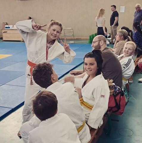 Junge Judoka in einem Dojo, lächelt und posiert, umgeben von trainierenden Kindern in weißen und gelben Judogis.
