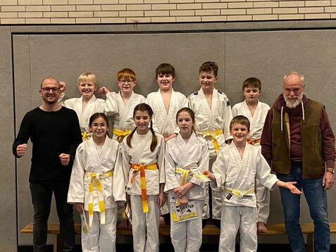 Gruppenzusammenstellung von zehn Kindern in Karateanzügen mit gelben und orangefarbenen Gürteln, zwei Erwachsenen stehen dabei.