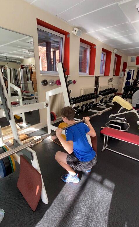 Junge mit blauer T-Shirt macht Kniebeugen mit Langhantel in einem Fitnessstudio. Gewichte und Spiegel im Hintergrund.