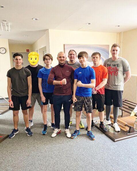 Gruppe von sechs Jugendlichen und einem Trainer in Sportkleidung, lächeln in einem Raum mit Fitnessgeräten.